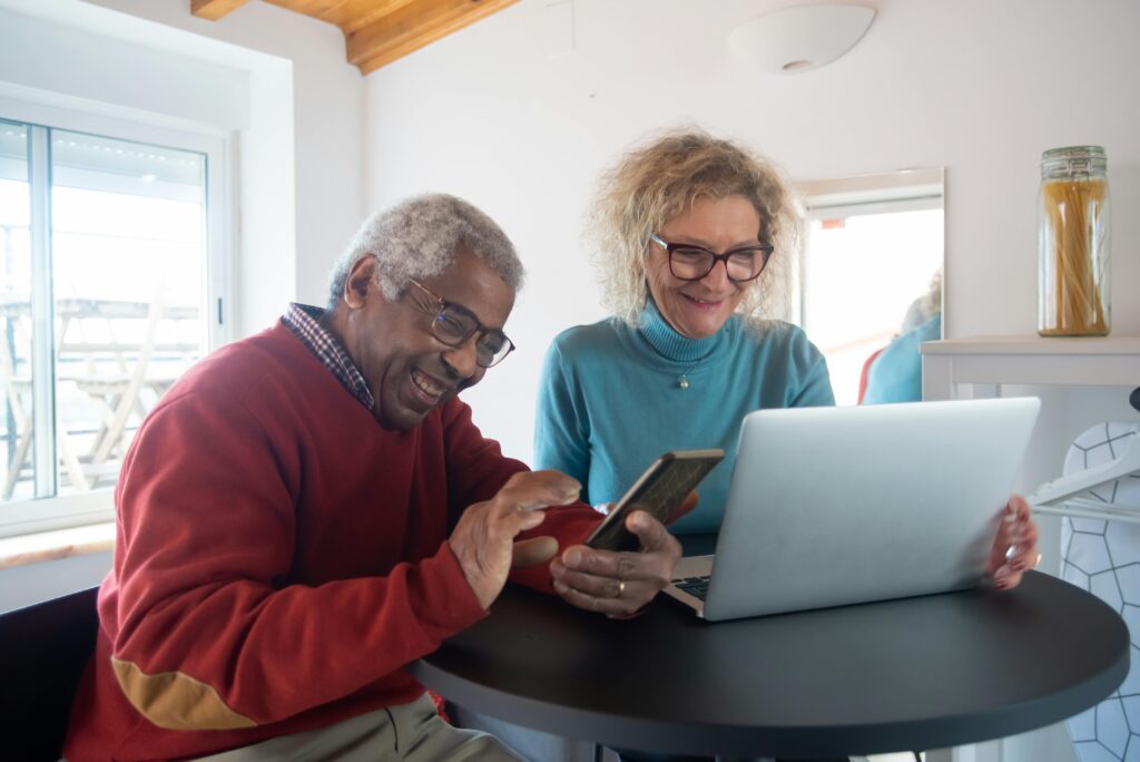 Smiling elderly couple using a smartphone and laptop indoors, enjoying online activities.
